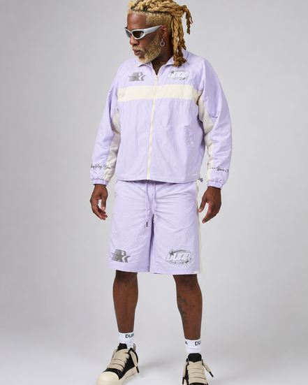 A model wearing a light purple and cream tracksuit consisting of a zip-up jacket and matching shorts, styled with white socks and beige-and-black sneakers against a neutral gray studio background.
