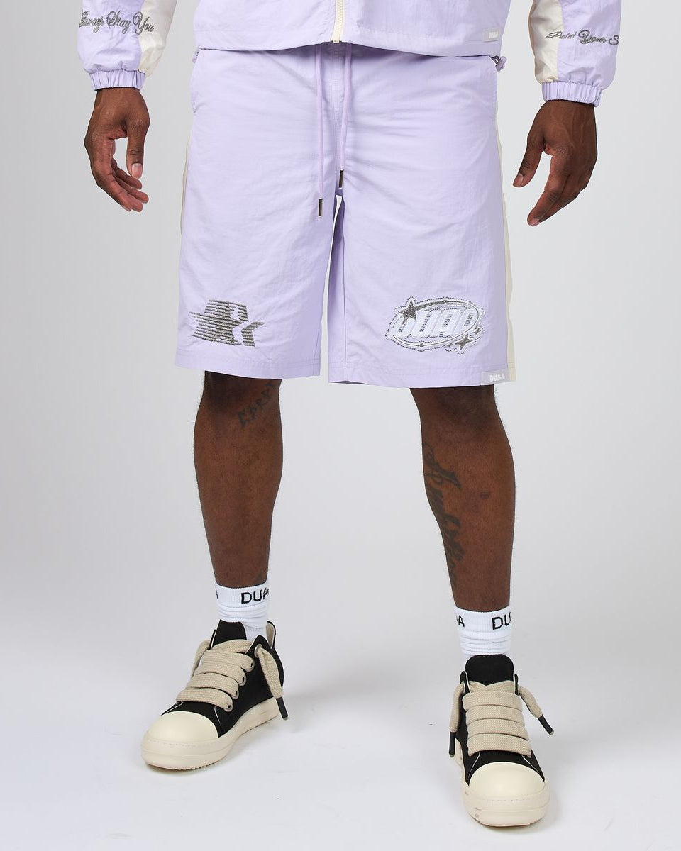 A person wearing light lavender Nike shorts with white side stripes and silver logo graphics, paired with white socks and beige-and-black sneakers, photographed against a light gray studio background.