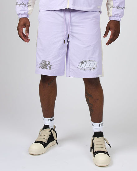 A person wearing light lavender Nike shorts with white side stripes and silver logo graphics, paired with white socks and beige-and-black sneakers, photographed against a light gray studio background.