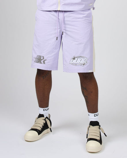 A person wearing light lavender athletic shorts with white socks and beige-and-black sneakers, shown from the waist down against a plain light gray studio background.