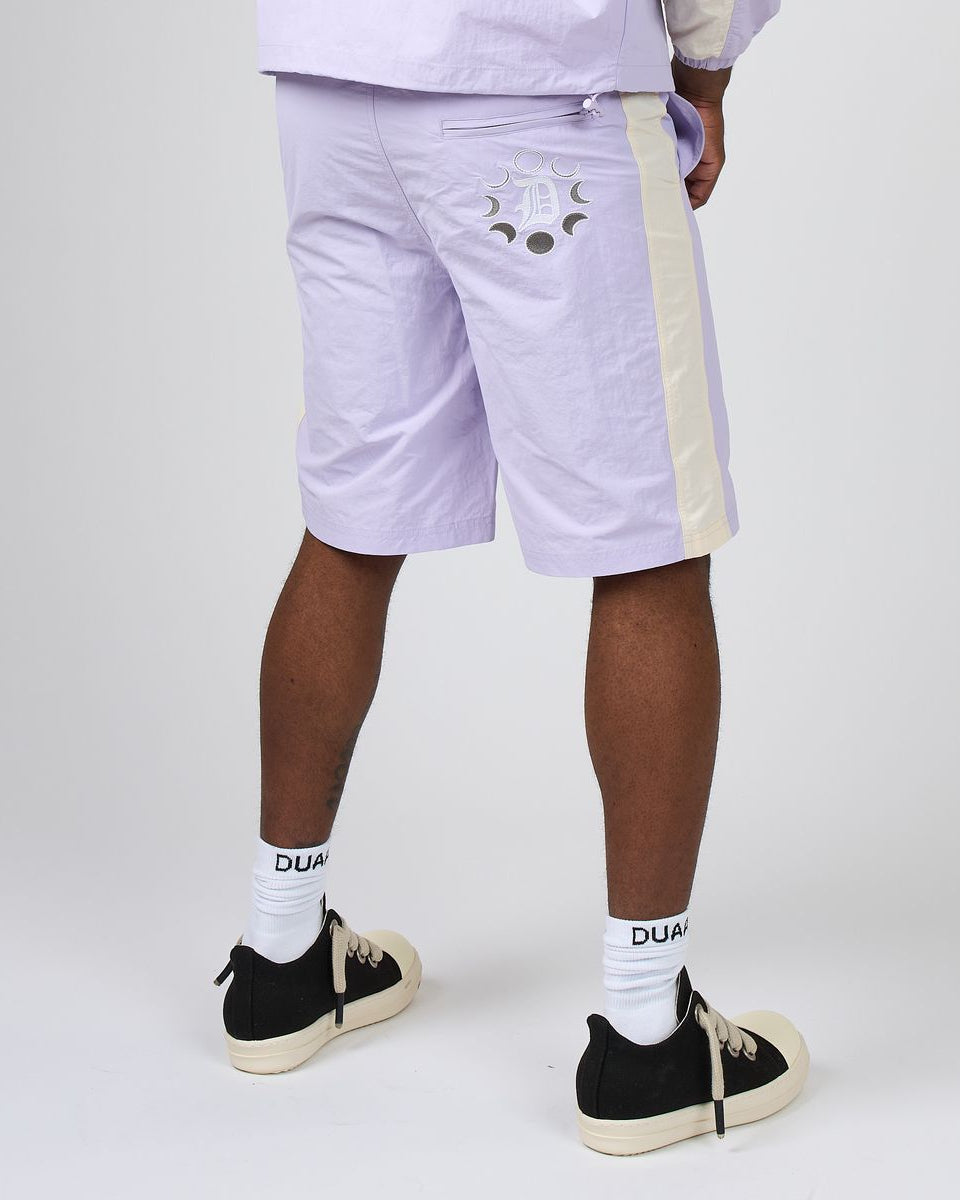 A person shown from the waist down wearing light lavender shorts with a cream side stripe and a moon graphic on the back pocket, paired with white socks and black sneakers, against a light gray studio background.