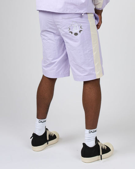 A person shown from the waist down wearing light lavender shorts with a cream side stripe and a moon graphic on the back pocket, paired with white socks and black sneakers, against a light gray studio background.