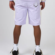 A person shown from the back wearing light lavender shorts with a small embroidered face detail on the left side, a matching light jacket, white socks, and black-and-white sneakers against a light gray studio background.