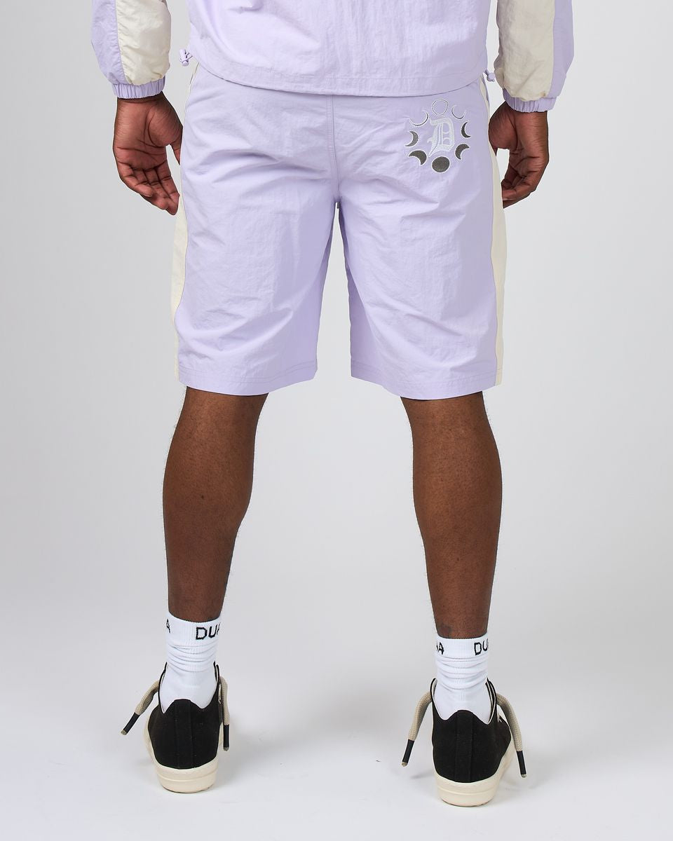 A person shown from the back wearing light lavender shorts with a small embroidered face detail on the left side, a matching light jacket, white socks, and black-and-white sneakers against a light gray studio background.