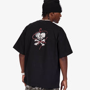 A man wearing a black oversized t-shirt with a skull and crossbones graphic on the back, paired with camouflage pants and a black beanie, shown from behind against a light gray background.
