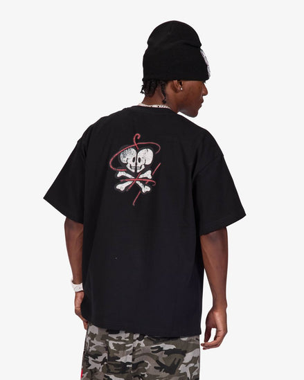 A man wearing a black oversized t-shirt with a skull and crossbones graphic on the back, paired with camouflage pants and a black beanie, shown from behind against a light gray background.