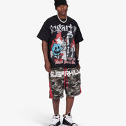 A man modeling a black graphic t-shirt, camouflage shorts with red side stripes, a black beanie, and black-and-white sneakers against a light gray studio background.
