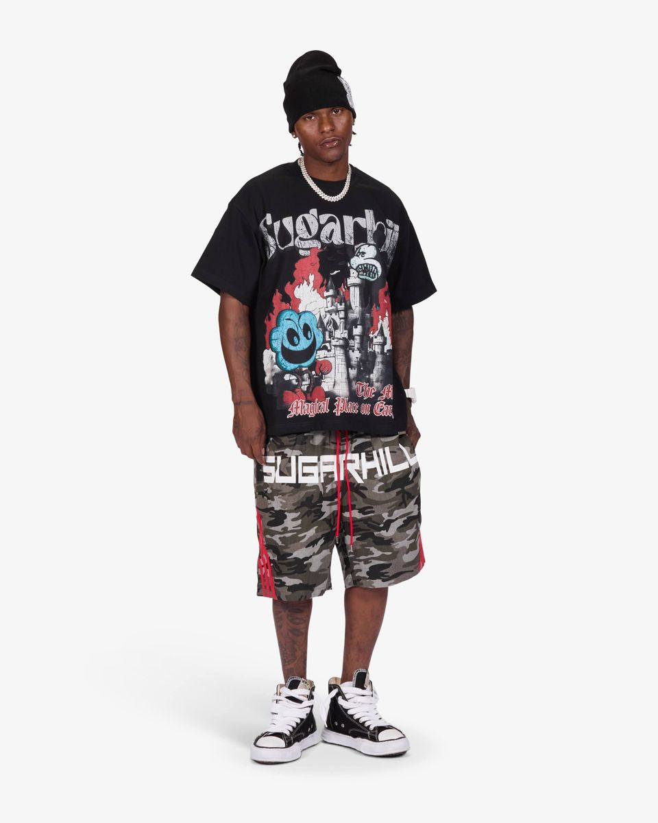 A man modeling a black graphic t-shirt, camouflage shorts with red side stripes, a black beanie, and black-and-white sneakers against a light gray studio background.