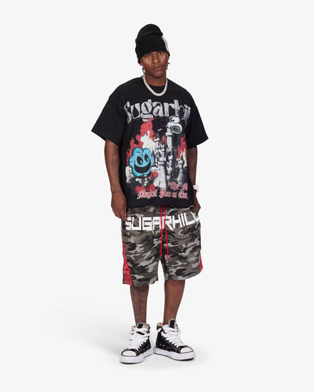 A man modeling a black graphic t-shirt, camouflage shorts with red side stripes, a black beanie, and black-and-white sneakers against a light gray studio background.