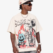 A man wearing a cream-colored graphic t-shirt with a Mickey Mouse and Donald Duck design, paired with a matching cap, sunglasses, and distressed jeans, photographed against a light gray background.