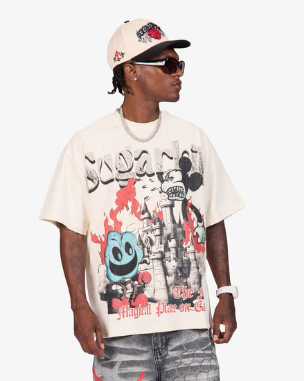 A man wearing a cream-colored graphic t-shirt with a Mickey Mouse and Donald Duck design, paired with a matching cap, sunglasses, and distressed jeans, photographed against a light gray background.