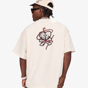 A person shown from the back wearing a cream-colored short-sleeve t-shirt with a red and black skull-and-crossbones graphic on the back, paired with a matching cream cap and dark sunglasses, against a light gray studio background.