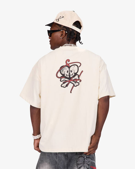 A person shown from the back wearing a cream-colored short-sleeve t-shirt with a red and black skull-and-crossbones graphic on the back, paired with a matching cream cap and dark sunglasses, against a light gray studio background.