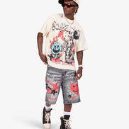 A model wearing a cream graphic t-shirt, gray denim shorts, and black high-top sneakers, standing against a light gray studio background.