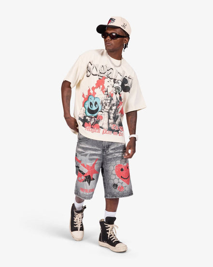 A model wearing a cream graphic t-shirt, gray denim shorts, and black high-top sneakers, standing against a light gray studio background.
