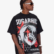 A man wearing a black oversized graphic t-shirt with a red and white design, paired with gray patterned jeans and sunglasses, photographed against a light gray background.