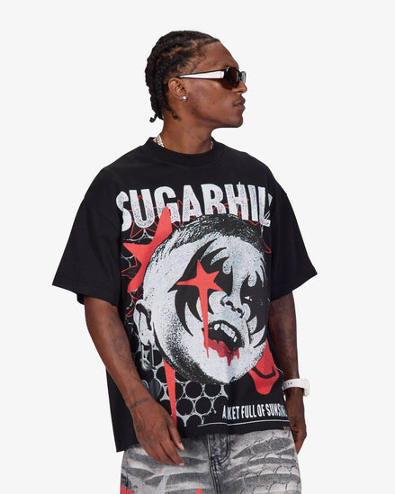 A man wearing a black oversized graphic t-shirt with a red and white design, paired with gray patterned jeans and sunglasses, photographed against a light gray background.