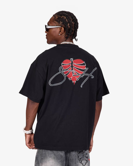 A man wearing a black oversized t-shirt from the back, featuring a red heart graphic with a basketball design and a white script logo. He is also wearing gray jeans, a silver chain necklace, and white-framed sunglasses against a light gray background.