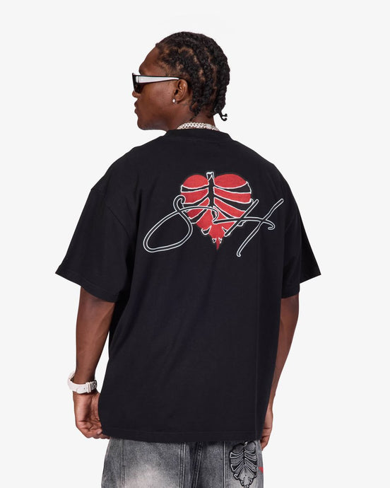 A man wearing a black oversized t-shirt from the back, featuring a red heart graphic with a basketball design and a white script logo. He is also wearing gray jeans, a silver chain necklace, and white-framed sunglasses against a light gray background.