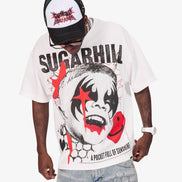 A man wearing a white oversized graphic t-shirt with a black and red clown face print and the text 'SUGARHILL' and 'A POCKET FULL OF SUNSHINE'. He is also wearing a black and white trucker hat and light-wash denim shorts against a plain white background.