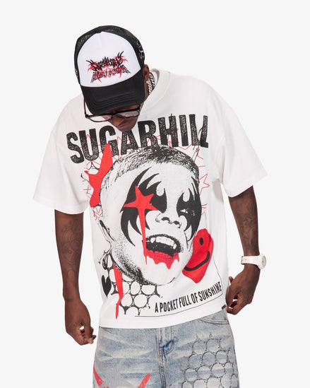 A man wearing a white oversized graphic t-shirt with a black and red clown face print and the text 'SUGARHILL' and 'A POCKET FULL OF SUNSHINE'. He is also wearing a black and white trucker hat and light-wash denim shorts against a plain white background.