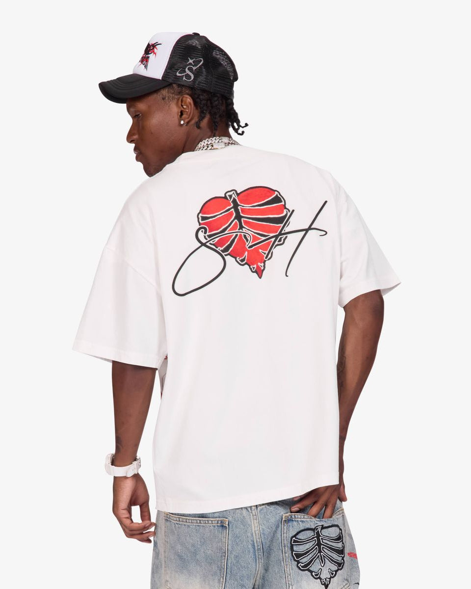 A man shown from the back wearing a white t-shirt with a red and black graphic on the back, paired with light-wash jeans and a black and white cap, against a light gray background.