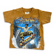 A vintage kids' t-shirt in a washed mustard brown color, featuring a large graphic print of a skateboarder in a city scene with the word 'GRINDING' in stylized lettering.