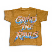 A short-sleeve crewneck t-shirt in a washed, mustard-brown color, featuring a large graphic print on the back that reads 'GRIND THE RAILS' in a stylized metallic font with blue, red, and silver colors.