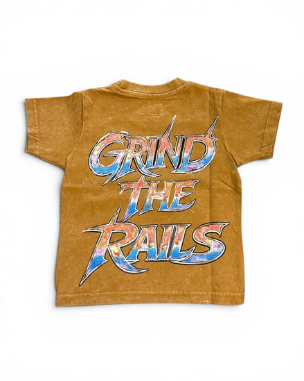 A short-sleeve crewneck t-shirt in a washed, mustard-brown color, featuring a large graphic print on the back that reads 'GRIND THE RAILS' in a stylized metallic font with blue, red, and silver colors.