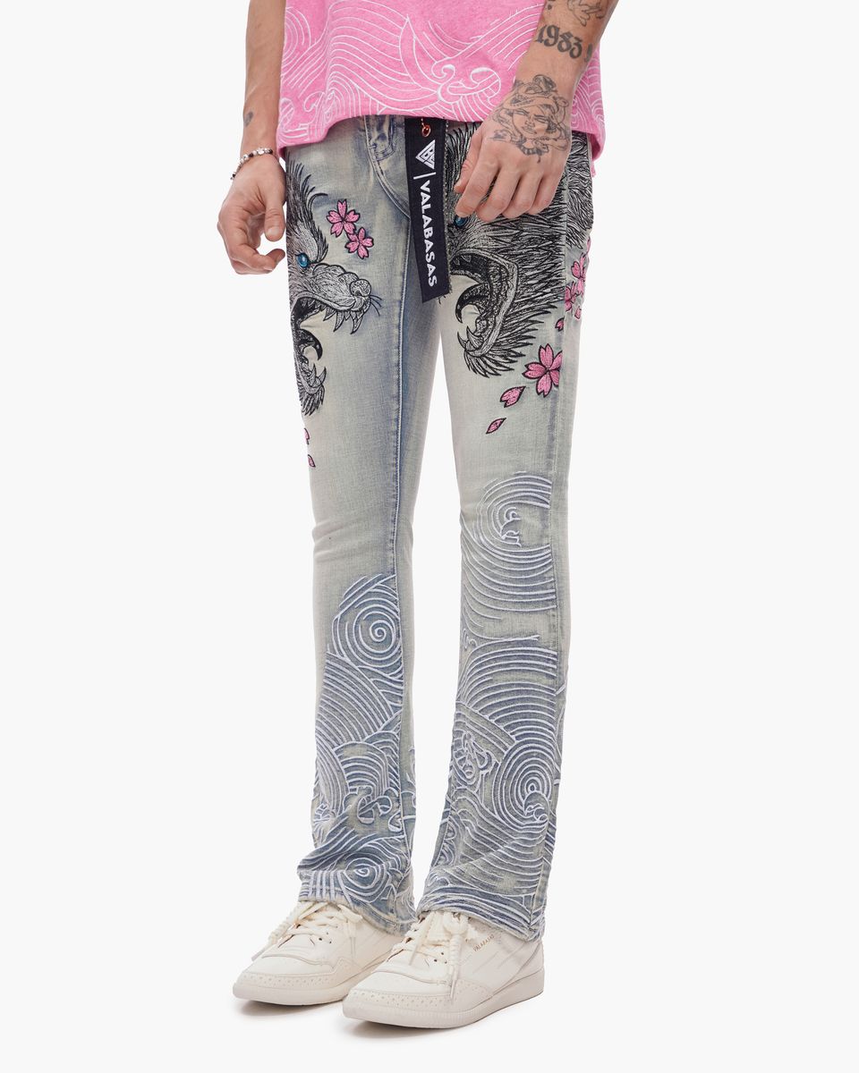Person wearing gray jeans with floral and abstract designs, white sneakers, and a pink top on a white background.