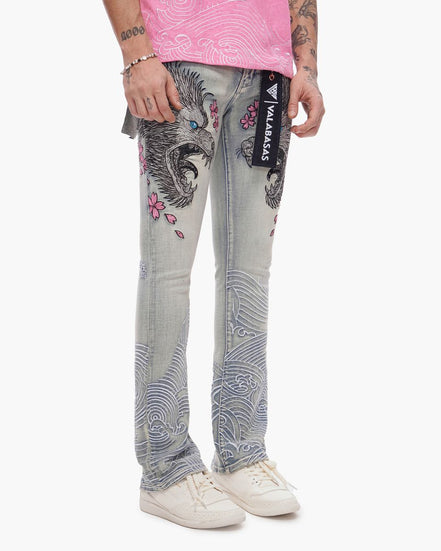 Person wearing blue jeans with floral and eagle design, pink shirt, and white sneakers on a white background