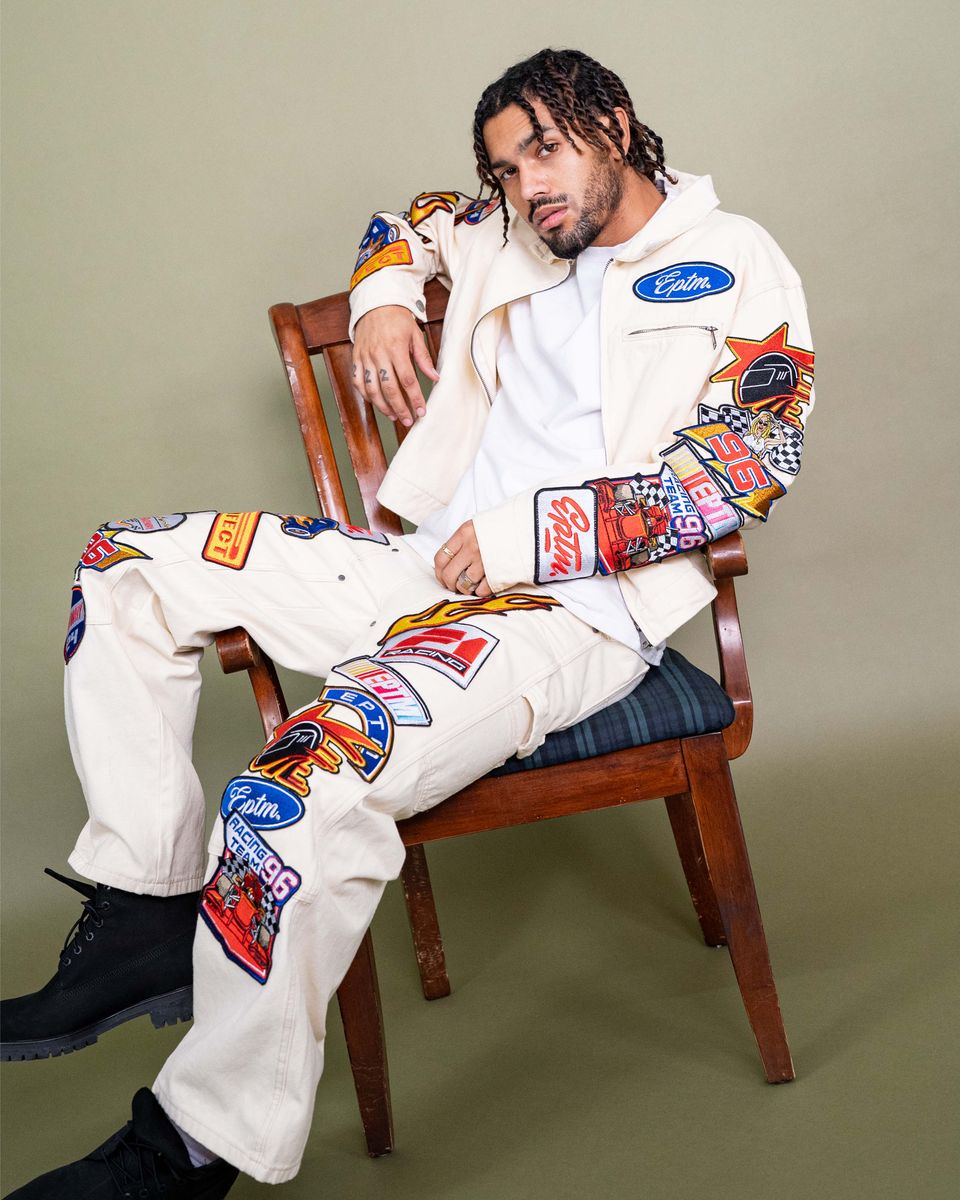 Person wearing a white outfit with colorful patches sitting on a wooden chair against a beige background