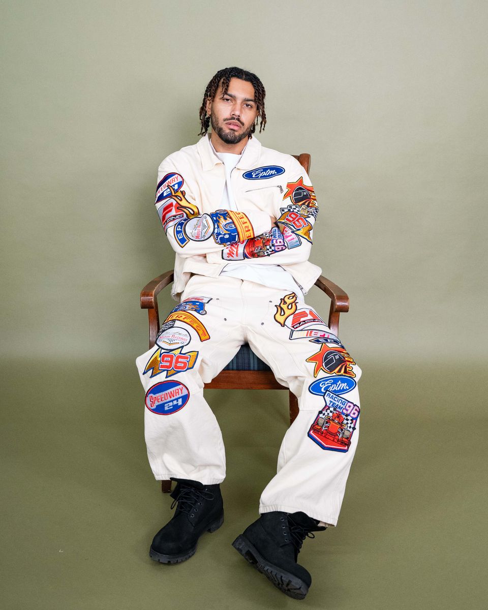 Person wearing a white jumpsuit with colorful patches sitting on a chair against a plain background