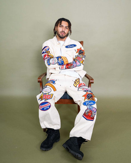 Person wearing a white jumpsuit with colorful patches sitting on a chair against a plain background