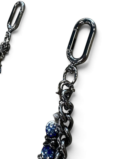 Close-up of a black metal chain accessory with a large carabiner-style clasp and dangling links, including two blue faceted beads near the bottom, on a white background.