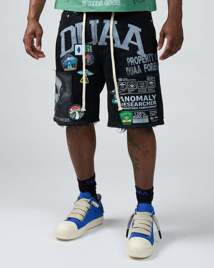 Person wearing black shorts with graphic designs and text, blue sneakers, and a green shirt on a white background.