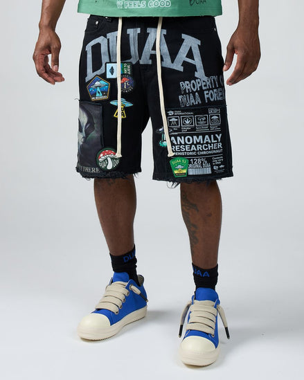 Person wearing black shorts with graphic designs and text, blue sneakers, and a green shirt on a white background.
