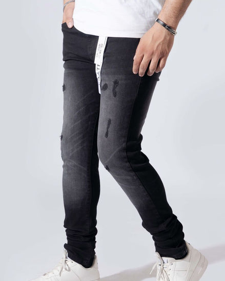 A person wearing slim-fit black distressed jeans with a white vertical stripe detail on the side, paired with a white t-shirt and white sneakers, shown from the waist down against a light gray studio background.