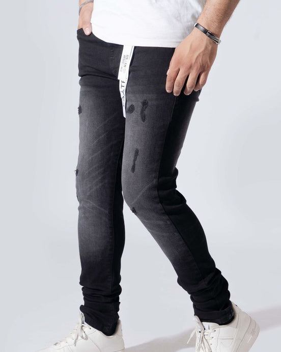 A person wearing slim-fit black distressed jeans with a white vertical stripe detail on the side, paired with a white t-shirt and white sneakers, shown from the waist down against a light gray studio background.