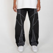 A person wearing black wide-leg pants with gray geometric paneling and white sneakers, shown from the waist down against a light gray studio background.