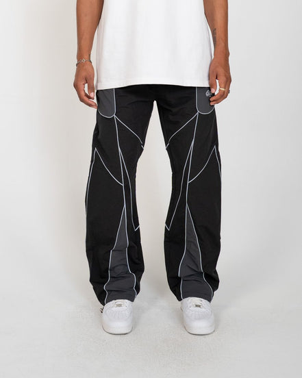 A person wearing black wide-leg pants with gray geometric paneling and white sneakers, shown from the waist down against a light gray studio background.