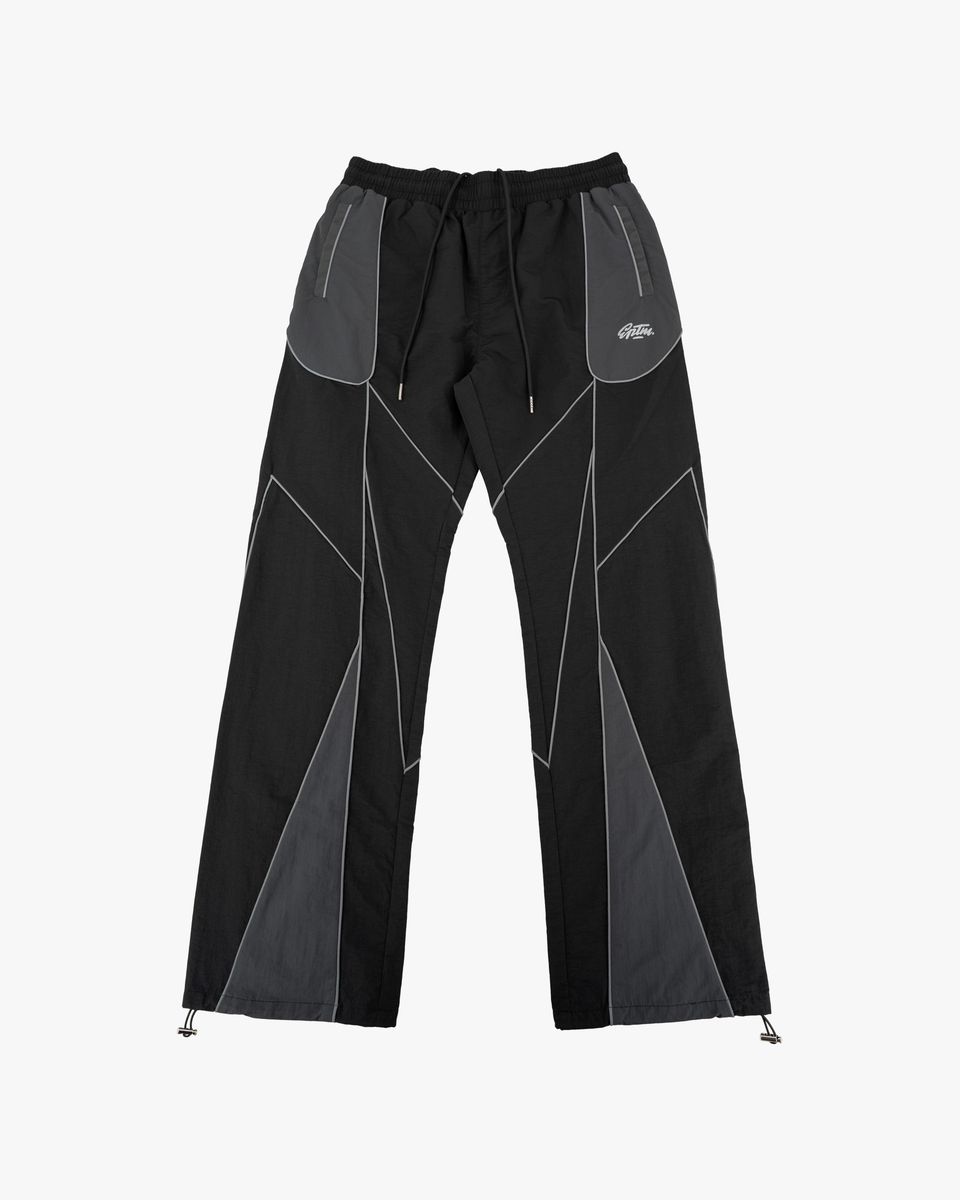 Black and gray geometric panel pants with a drawstring waist and zippered side pockets, shown flat against a light gray background.