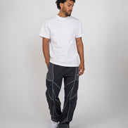 A man modeling a white short-sleeve crewneck t-shirt and dark gray pants with white geometric contrast stitching, standing against a light gray studio background.