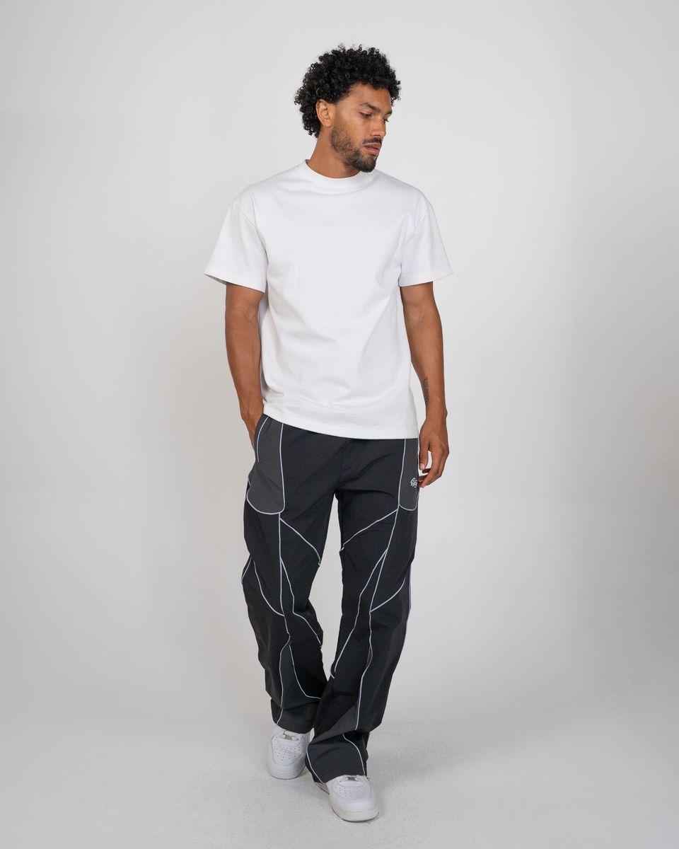 A man modeling a white short-sleeve crewneck t-shirt and dark gray pants with white geometric contrast stitching, standing against a light gray studio background.