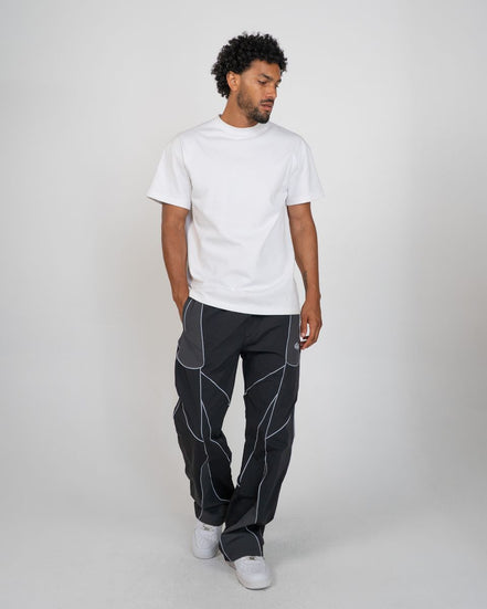A man modeling a white short-sleeve crewneck t-shirt and dark gray pants with white geometric contrast stitching, standing against a light gray studio background.
