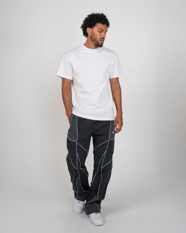 A man modeling a white short-sleeve crewneck t-shirt and dark gray pants with white geometric contrast stitching, standing against a light gray studio background.