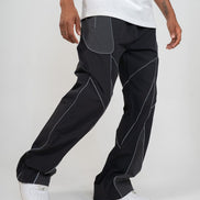 A person wearing loose black pants with contrasting light gray paneling and white sneakers, cropped from the waist down against a plain light gray studio background.