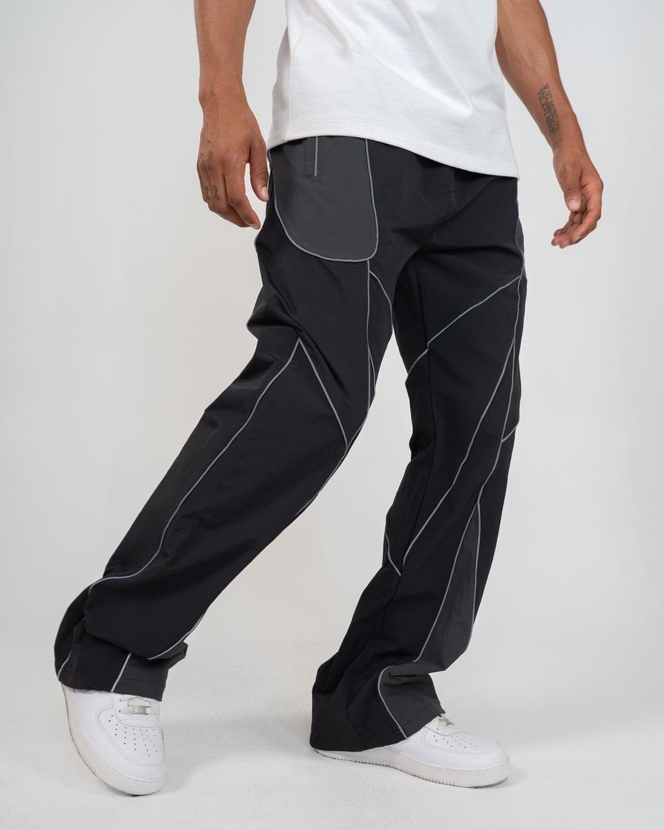 A person wearing loose black pants with contrasting light gray paneling and white sneakers, cropped from the waist down against a plain light gray studio background.