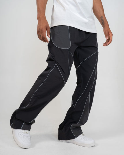 A person wearing loose black pants with contrasting light gray paneling and white sneakers, cropped from the waist down against a plain light gray studio background.