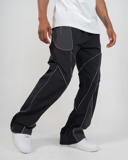 A person wearing loose black pants with contrasting light gray paneling and white sneakers, cropped from the waist down against a plain light gray studio background.